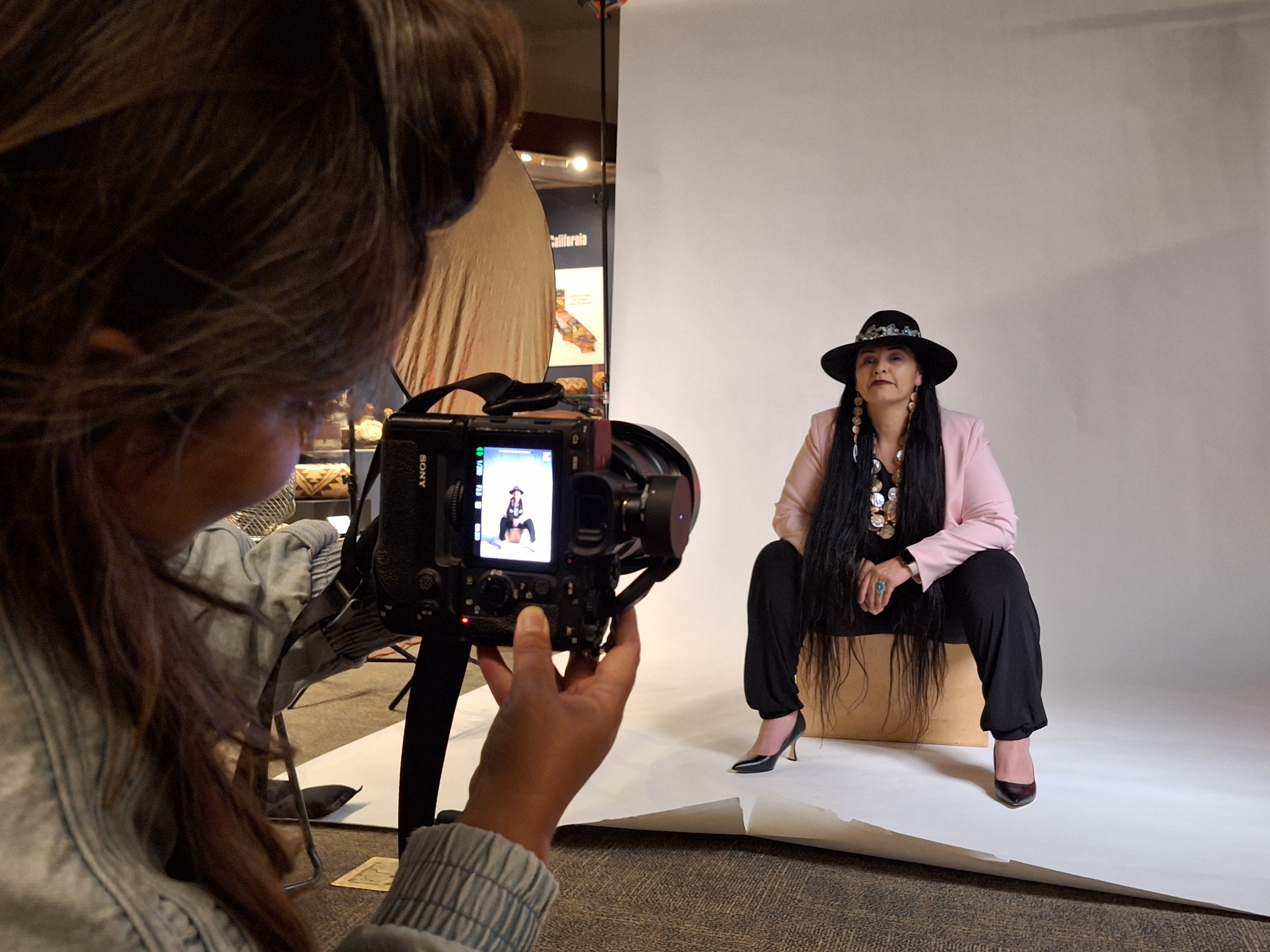 A woman in a hat with very long hair is being photographed and is visible in the view finder of the photographer's camera. 