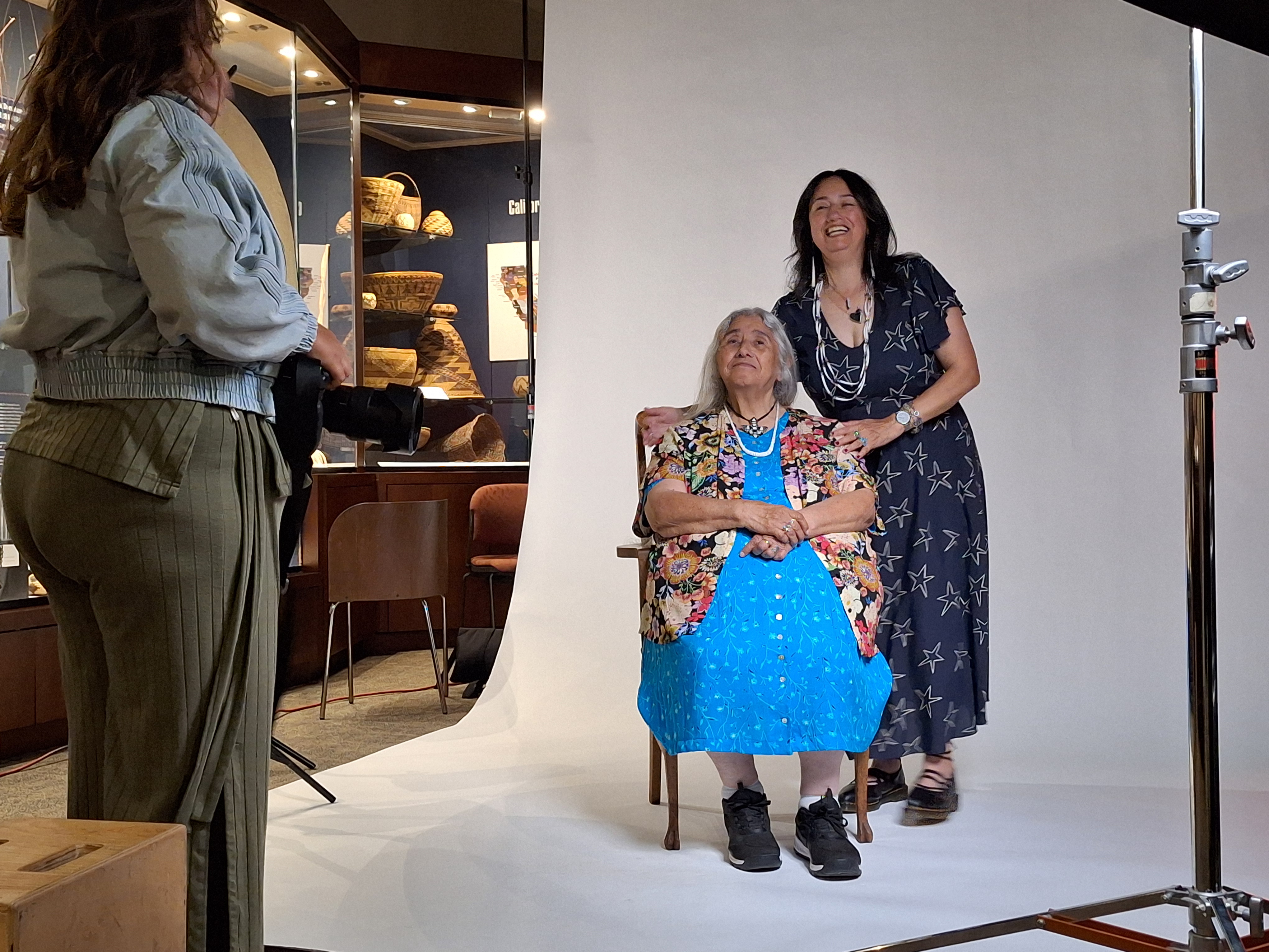 A woman stands next to her elder mother who is seated on a chair next to her, ready to be photographed.